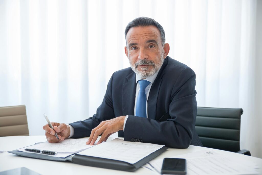 A bearded man in black suit sitting near the table with documents